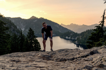 Man on ridge at sunset over lake view resting with hands on knees.