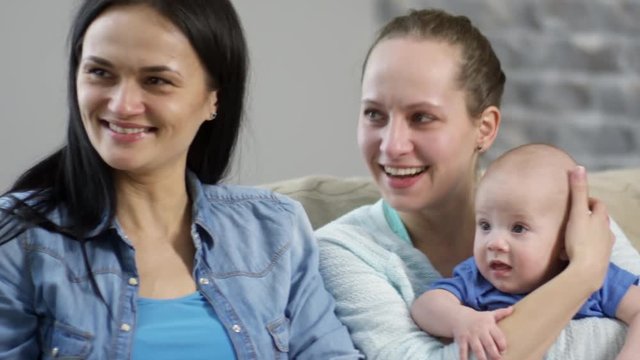 Handheld medium shot of happy female couple sitting on sofa and laughing while spending time with cute baby boy