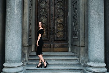 Cute young girl modeling appearance posing against the backdrop of huge beautiful doors in black dress and hat