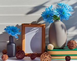 An empty wooden frame is on a white wooden background. Nearby is a gray vase with blue chrysanthemums or daisies on a stack of books. Around are scattered wicker balls.