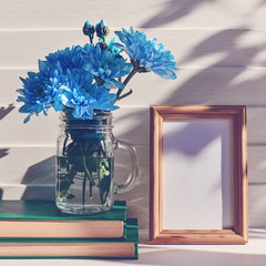 An empty wooden frame is on a white wooden background. Nearby is a gray vase with blue chrysanthemums or daisies on a pile of books.