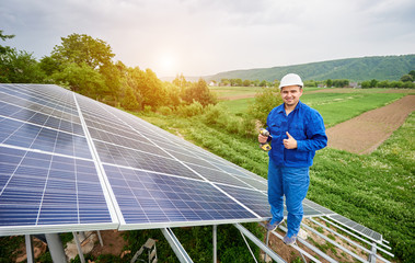 Construction worker with screwdriver looking in camera with thumb-up gesture on photo voltaic panel solar system shiny surface and lit by sun green fields background. Alternative energy concept.