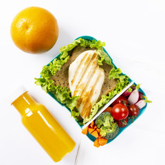 Lunch box with a useful and tasty meal. Grilled chicken, vegetables, orange and a bottle of orange juice. The background is white. The concept of a healthy lifestyle. Top view.