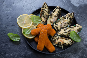 Baked mussels and fried fish sticks or breaded fish fillet served on a metal tray over grey concrete background