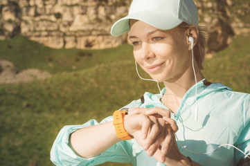 Portrait of a smiling young fitness girl in a cap and headphones checking her smart clock while sitting outdoors against a background of rocks