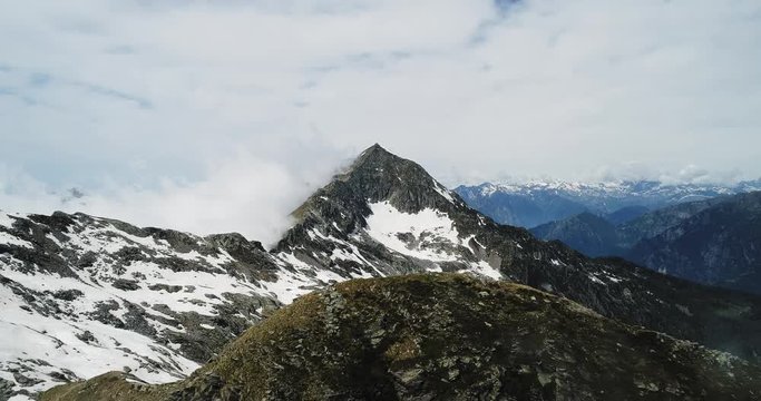Backward aerial top view over cloudy rocky snowy mountain in sunny day with clouds.Italian alps mountains in summer with wild windy weather outdoor nature establisher.4k drone flight establishing shot