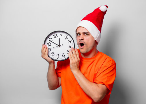 Young handsome man in orange t-shirt with Christmas hat and big clock. Studio image on white background