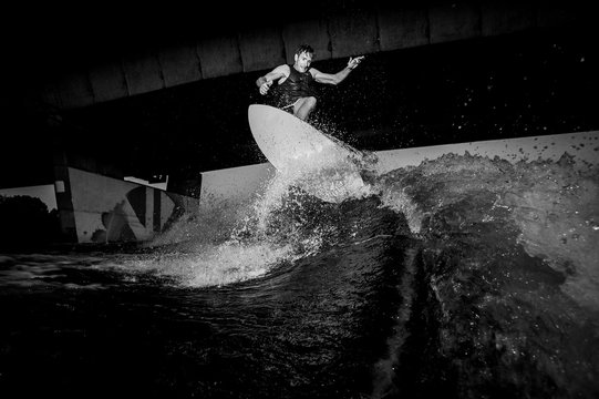 Black and white photo of active wakesurfer riding on river waves against the bridge