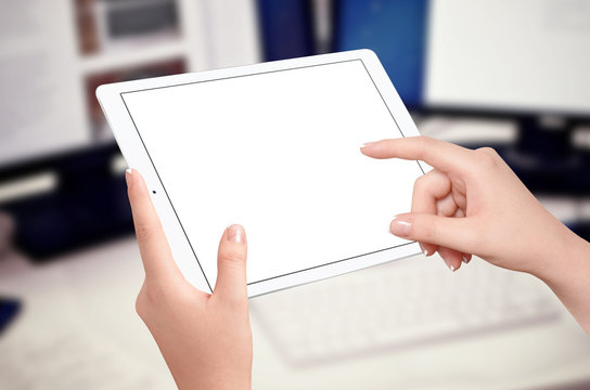 Female hands holding and touching empty screen of white tablet in horizontal position, blurred office desk with computer in background. Mockup