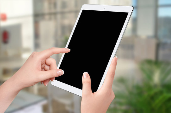 Female hands holding and touching empty screen of white tablet in vertical position, blurred office room in background. Mockup