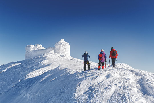 Three brave rescuers explore the terrain of snowy slope for avalanche danger against snow capped old observatory at peak of Carpathian mountain. Rear view. Winter seasonal landscape.