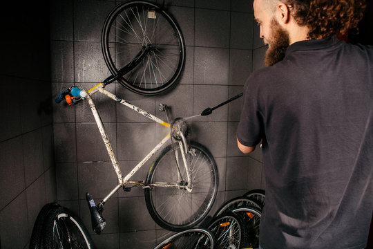 Services professional washing of a bicycle in the workshop. Close-up of hand Young Caucasian stylish man doing bicycle cleaning using automatic electric water pump. Sprays scatter from the pressure