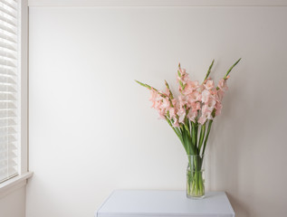 Pink gladioli flowers in glass vase on white table against wall next to window.