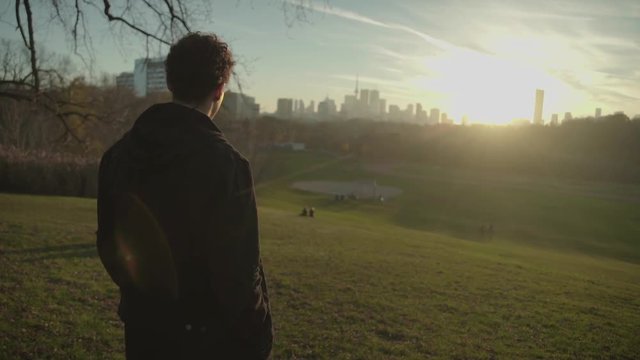 Confident Young Adult Walks to Bottom of Hill and Stops in Urban Park to Look at City, Cinematic Slow Motion Gimbal Shot with Golden Hour Lens Flares at Sunset