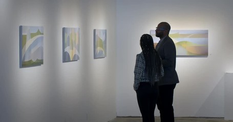 MLS African American man and Hispanic woman talking and looking at abstract paintings in art gallery. Wide view with couple in profile - Powered by Adobe
