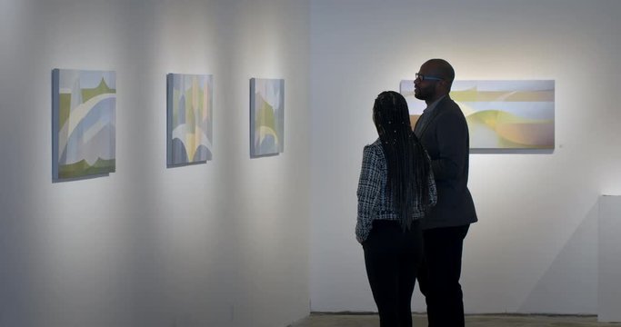 MLS African American man and Hispanic woman talking and looking at abstract paintings in art gallery. Wide view with couple in profile