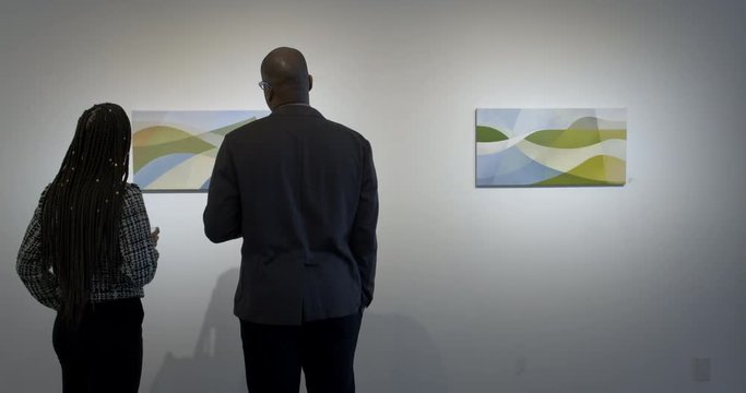 MS African American man and Hispanic woman with long braids look at paintings in gallery during art show opening reception. Locked off, couple enter shot and stand with backs to camera