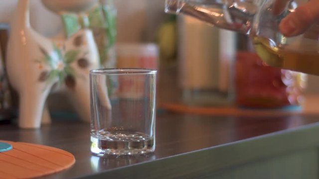 Hand pouring bourbon whiskey from a curved bottle into a clear glass sitting on a dark wood home bar with kick-knacks and other bottles in the background