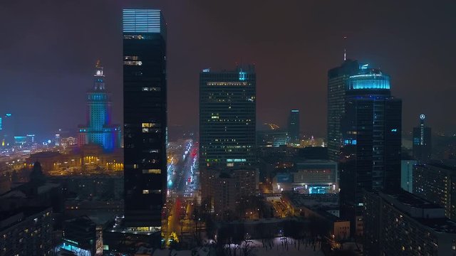 Drone footage of warsaw center at night. In the front there are modern, glazed skyscrapers, in the background the Palace of Culture and Science.