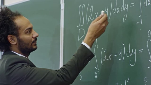 Handheld shot of friendly male professor standing at blackboard and explaining equations written on it, then turning to students and asking questions