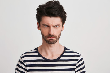 Closeup of mad irritated young man with bristle wears striped t shirt feels angry and looks directly in camera isolated over white background