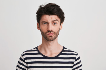 Portrait of pensive confused young man with bristle wears striped t shirt feels embarrassed and thinking isolated over white background Looks directly in camera