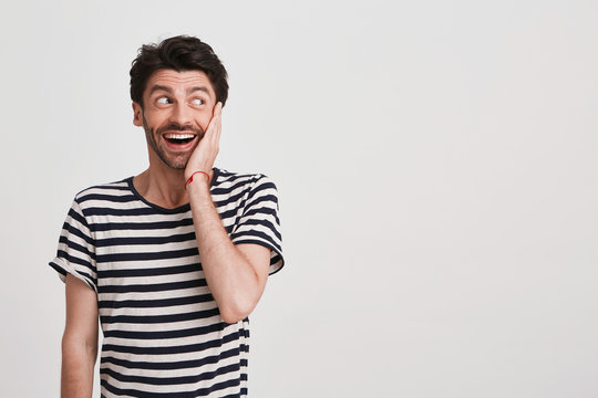 Closeup of surprised excited young man with bristle and hand on cheek wears striped t shirt feels happy and looks to the side isolated over white background