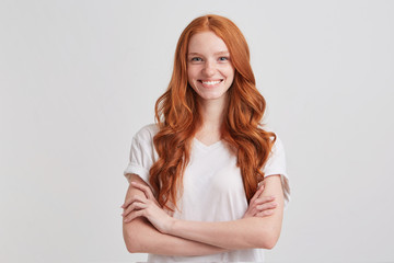 Closeup of smiling lovely redhead young woman with long wavy hair and freckles wears stylish t shirt looks confident and keeps arms crossed isolated over white background
