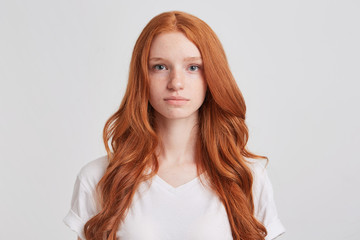 Closeup of serious beautiful redhead young woman with long wavy hair and freckles wears t shirt feels confident and looks directly in camera isolated over white background - obrazy, fototapety, plakaty