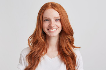 Closeup of cheerful lovely redhead young woman with long wavy hair and freckles wears t shirt looks confident and feels happy isolated over white background Looks directly in camera