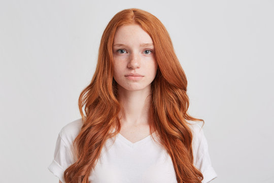 Closeup of serious beautiful redhead young woman with long wavy hair and freckles wears t shirt feels confident and looks directly in camera isolated over white background