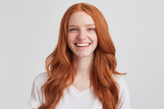 Portrait of cheerful pretty redhead young woman with long wavy hair and freckles wears t shirt feels joyful and laughing isolated over white background Looks directly in camera