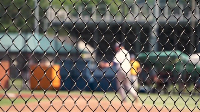 A pitcher throws a pitch and gives up a base hit in a youth baseball game. Game is viewed through a chain link fence, with selective focus on the fence and the players and game action out of focus