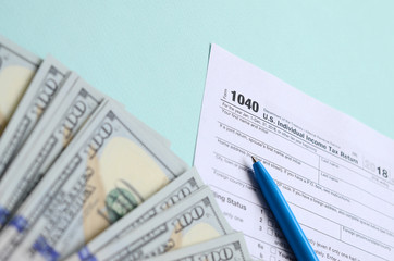 1040 tax form lies near hundred dollar bills and blue pen on a light blue background. US Individual...
