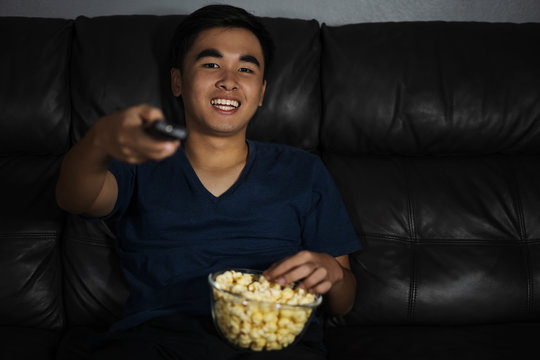 cheerful man holding remote control and watching TV while sitting on sofa at night