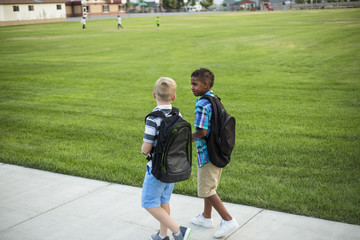 Naklejka premium Two diverse school kids walking and talking together on the way to school. Back to school photo of diverse school children wearing backpacks in the school yard