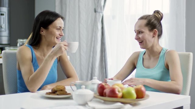 PAN of happy young female couple chatting and drinking tea in kitchen