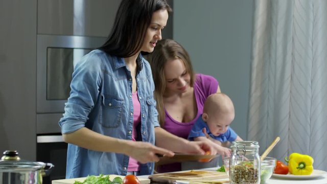 Tilt up of young woman cutting vegetables in kitchen and chatting with female partner holding curious little boy nibbling leaf of lettuce, then wiping her hands on towel and interacting with baby