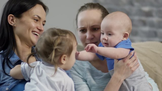 Medium shot of cute toddler girl and baby boy sitting on arms of happy mothers and interacting with each other