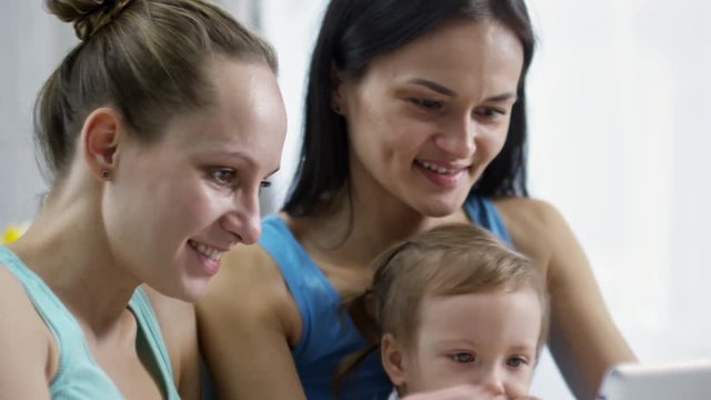 Tilt up of cheerful same-sex female couple with toddler girl watching cartoons on tablet
