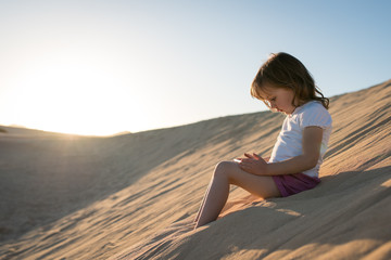 Fototapeta na wymiar Little girl sitting on sand dune and playing game on phone.