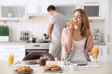 Beautiful young woman having breakfast while her beloved boyfriend cooking in kitchen - obrazy, fototapety, plakaty