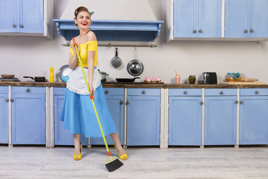 Retro / pin up girl woman female / housewife wearing colorful top, skirt and white apron holding mop and cleaning washing floor in the kitchen with blue cabinets and utensils. Housework concept