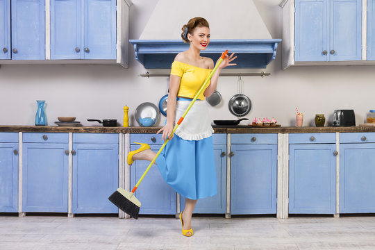 Retro / pin up girl female woman / housewife wearing colorful top, skirt and white apron holding mop and cleaning washing floor in the kitchen with blue cabinets and utensils. Housework concept
