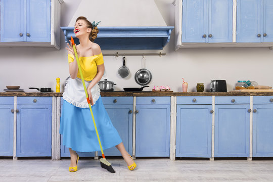 Retro / pin up girl woman female / housewife wearing colorful top, skirt and white apron holding mop singing and cleaning floor in the kitchen with blue cabinets and utensils. Housework concept