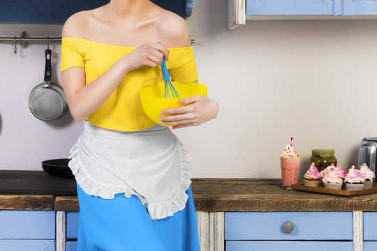 Retro beauty pin up girl housewife wearing colorful top, skirt and white apron holding whipper and bowl making sweets in the kitchen with blue cabinets and utensils and cupcakes with milkshake. 