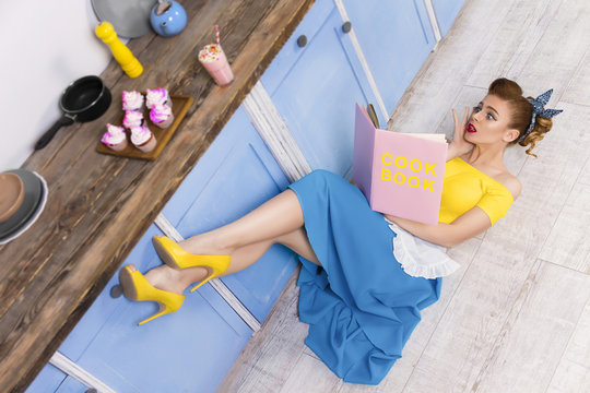 Colorful retro / pin up girl / housewife wearing colorful top, skirt and white apron lying on the floor in the kitchen and reading pink book. Sweet food cupcakes and milkshake home cooked on the table
