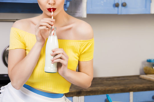 Colorful retro pin up girl woman female housewife wearing colorful top, skirt and white apron drinking milk from glass of bottle sitting in the kitchen. Retro styled