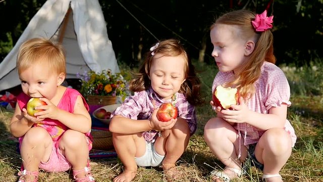 Three little cute child girls funny eat red apples on nature picnic in slow motion