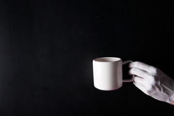 White painted hand holding a white cup on a black background.  Abstract. Copy space.